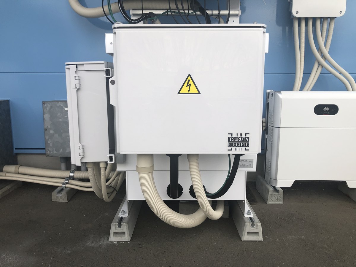 実際に設置された絶縁トランスの写真。白い金属製の箱の扉に高電圧注意の表示があり、下部から複数の配管が接続されている。左に小型の盤、右に別の機器が並んで設置されている